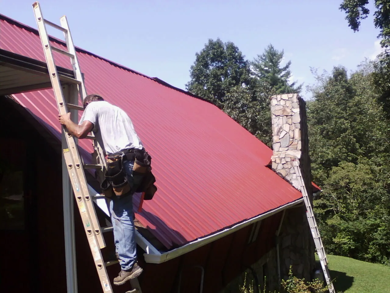 Roofer on a ladder beside a red metal roof and stone chimney — Soffit & Fascia Repair by Roofing Brothers in Granbury
